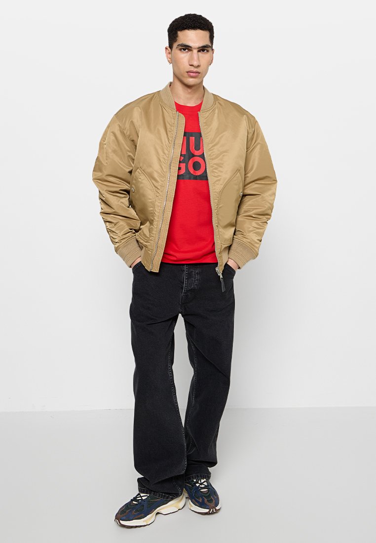 Chaqueta bomber color marrón con puños acanalados, llevada sobre una camiseta gráfica roja, combinada con jeans negros y zapatillas multicolores. Look simple y casual.