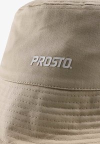 Beżowa capka o kształcie wiadra, wykonana z teksturowanej tkaniny, z haftowanym wzorem oraz białym logo "PROSTO." z przodu.