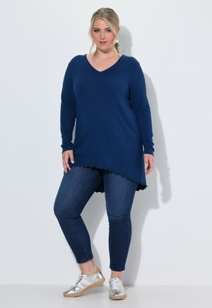 Vrouw draagt een marineblauwe asymmetrische trui, donkerblauwe skinny jeans, zilveren sneakers en zwarte druppelvormige oorbellen, staand tegen een effen muur.