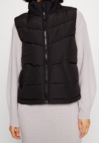 Černá vatovaná vesta s vysokým límcem, předním zipem a bočními kapsami. Má prošívaný vzor a volnější střih pro pohodlí.
