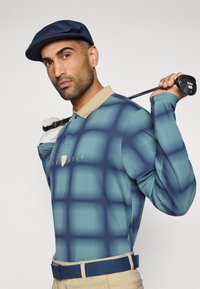 Μακρυμάνικο πόλο με διαβαθμισμένο μπλε σχέδιο, μπεζ γιακά και λογότυπο. Το μοντέλο κρατάει μπαστούνι γκολφ, φοράει ναυτικό καπέλο και μπεζ παντελόνια με μπλε ζώνη.