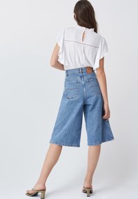 Culottes en denim taille haute de couleur bleu clair, avec un design à jambe large, dotés de poches arrière et d'un patch en cuir marron de la marque. Blouse blanche avec des manches à volants.