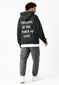 Felpa nera con cappuccio con testo bianco sul retro che recita "CREDO NEL POTERE DELL'AMORE", abbinata a pantaloni cargo grigi e scarpe da ginnastica bianche.