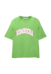 T-shirt in cotone verde con una vestibilità rilassata, scollatura rotonda e maniche corte. Presenta la scritta "KOMBUCHA" stampata in lettere rosa bold sulla parte anteriore.