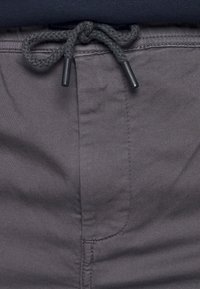 Grå tygshorts med en slät textur. Har en dragsnörning i midjan med svarta spetsar och sidofickor. Närbild.