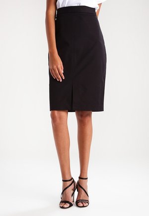 Vrouw draagt een zwarte kokerrok tot op de knie met een voorzak, een wit bovenstuk en zwarte sandalen met hoge hakken en bandjes, tegen een witte achtergrond.