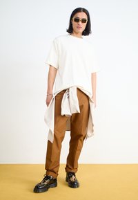 Witte oversized katoenen t-shirt, bruine broek, gelaagde beige shirt geknoopt rond de taille, en zwarte schoenen met een patroon. Gele zonnebril.