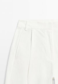 Close-up of white pants showing waistband, belt loops, side pocket, and front pleat detail on a plain background.