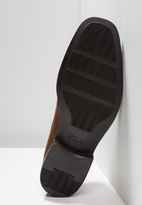 Hnědá kožená podrážka s texturovanou černou gumovou podrážkou s vzorem herringbone. Název značky "Clarks" je vyražen na patě.