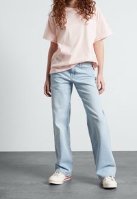 Osoba oblečená v světle růžovém volném tričku, světle modrých širokých džínách a bílých teniskách stojící na šedé podlaze.