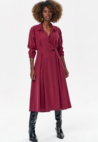 Kobieta z kręconymi włosami, ubrana w bordową sukienkę kopertową z długim rękawem i czarne, sięgające do kolan kozaki, stojąca na białym tle.