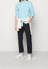 Světle modrá pruhovaná košile s knoflíky, tmavé džíny, krémový svetr svázaný kolem pasu, bílé tenisky, volný střih, minimalistický design.