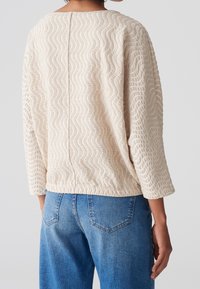 Haut beige texturé avec motif ondulé, manches trois-quarts et coupe décontractée, associé à un jean bleu avec poches arrière.