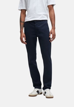 Uomo che indossa pantaloni dritti blu scuro, scarpe da ginnastica bianche e nere e una camicia bianca a maniche corte con tatuaggi visibili sulle braccia.