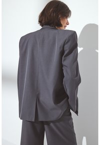 Blazer grigio su misura con una vestibilità rilassata, spalle prominenti e una singola fessura sul retro. Tessuto liscio con una leggera lucentezza.