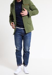 Grön lättviktig jacka med framfickor, bärs över en marinblå tröja, i kombination med blå jeans och vita sneakers, vilket visar en avslappnad stil.