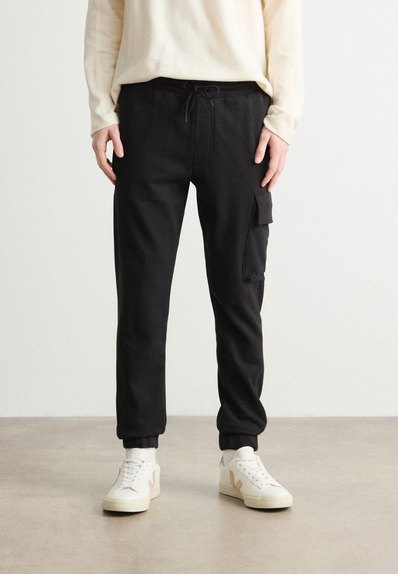 Pantalones de chándal negros con cintura ajustable, bolsillo lateral tipo cargo y puños ajustados. Llevados con zapatillas blancas y un suéter beige.
