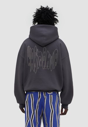 Person mit dunklem lockigem Haar trägt einen dunklen oversized Hoodie mit verzerrtem Text und blau gestreifter Hose, blickt von hinten vor einem einfarbigen Hintergrund.
