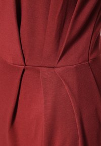 Gros plan sur une robe bordeaux, mettant en avant un tissu doux, des sections froncées et une texture lisse. Présente des coutures marquées et des plis nets.