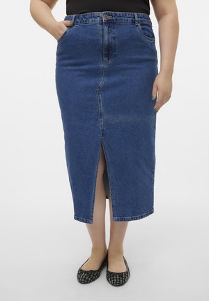 Kvinde iført en blå midi denim nederdel med en frontslids, sort top og sorte mønstrede ballerinaer, stående mod en hvid baggrund.