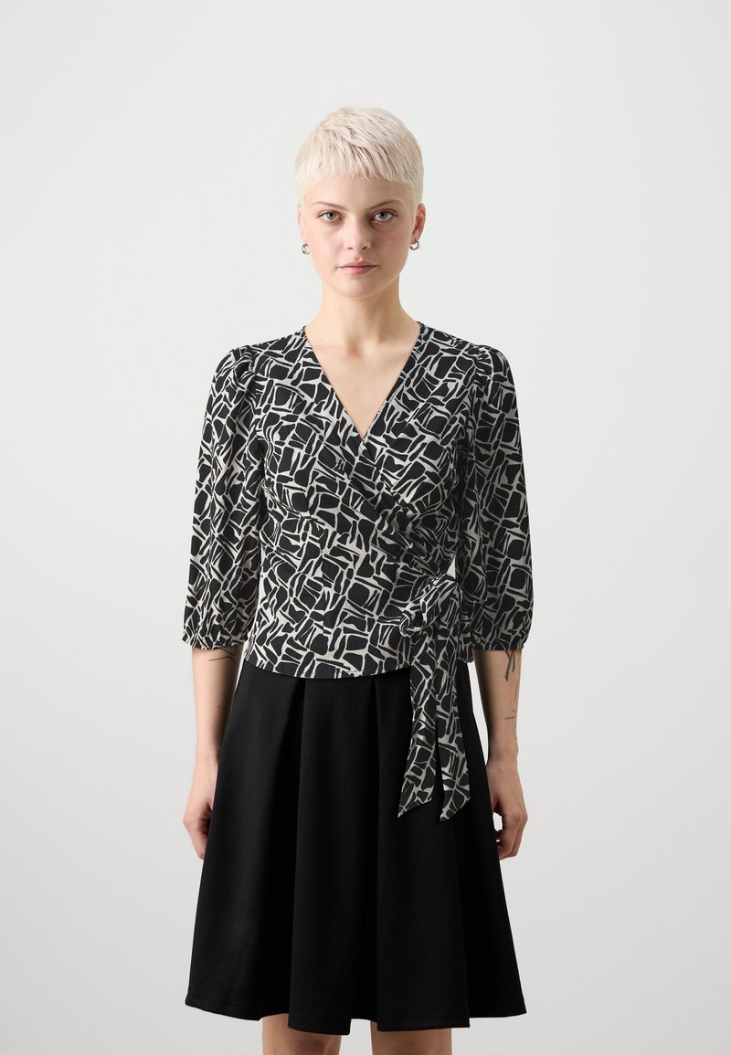 Schwarz-weiße Wickelbluse mit Dreiviertelärmeln, geometrischem Muster und seitlichem Bindeband, kombiniert mit einem schwarzen Schlagrock.
