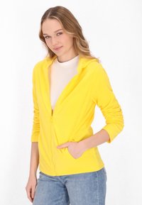 Felpa gialla con zip e maniche lunghe, realizzata in materiale morbido. Presenta tasche e una vestibilità comoda. Indossata sopra una maglietta bianca e abbinata a jeans.
