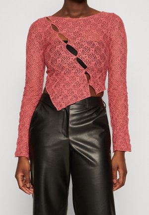 Różowy koronkowy crop top z długim rękawem o ukośnym wzorze wycięć, połączony z czarnymi, skórzanymi spodniami z wysokim stanem na stojącej osobie.