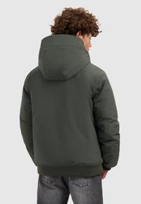 Jongere met krullend haar, gekleed in een donkergroene hoodie en vervaagde zwarte spijkerbroek, met de rug naar de camera gericht tegen een effen achtergrond.