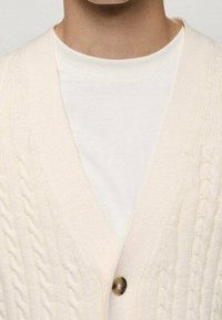 Cream cable-knit cardigan with a V-neck design and single button closure, layered over a plain white T-shirt. Soft texture, relaxed fit.