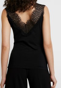 Svart ärmlös topp med en djup spets V-ringning på baksidan och fransade kanter, tillverkad av mjukt bomullsmaterial.