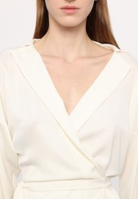 Cream wrap blouse with a V-neck and long sleeves, featuring a smooth, soft texture and a tie detail at the waist.