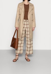 Femme portant un trench-coat beige, une chemise marron, un pantalon court à carreaux beige, des sandales marron, tenant un sac fourre-tout en cuir marron, debout sur un sol gris.