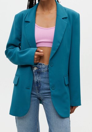 Personne portant un blazer bleu sarcelle par-dessus un crop top rose avec un jean bleu taille haute, tenant le revers du blazer d'une main.