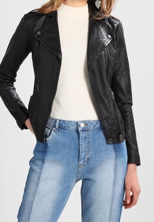 Femme portant une veste en cuir noire par-dessus un pull blanc côtelé avec un jean bleu taille haute, debout avec une main dans la poche sur fond gris.