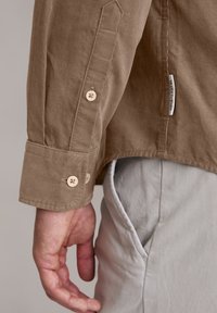 Brązowa koszula sztruksoowa z zapięciem na guziki, posiadająca jasne guziki. Widoczna metka z boku, połączona z jasnoszarymi spodniami.