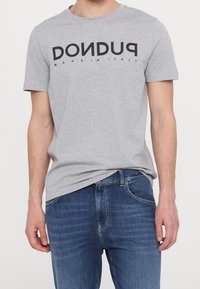 Camiseta de algodón gris con texto en negrita negro "DON DUD" en el frente. Manga corta, cuello redondo y ajuste relajado. Combinada con vaqueros azul oscuro.