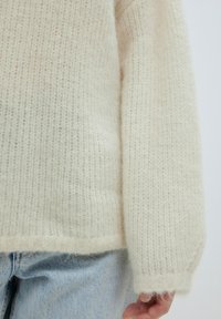 Cremfarbener Strickpullover mit einer weichen, flauschigen Textur, breiten Ärmeln und einem lockeren Schnitt, kombiniert mit hellblauen Jeans.