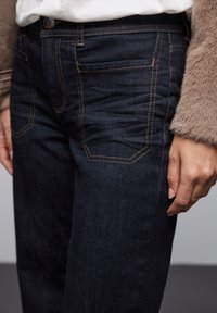 Jeans en denim bleu foncé avec des coutures visibles, présentant une coupe slim et des poches avant. Le tissu a une texture lisse. Design de jambe fuselée.