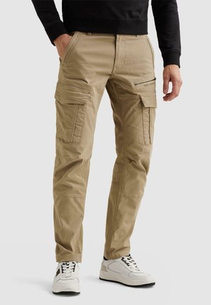 Man die beige cargobroek met meerdere zakken en witte sneakers draagt, staand met één hand in zijn zak tegen een effen achtergrond.