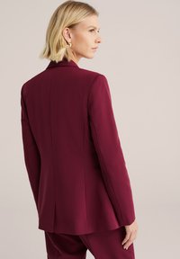 Burgundský přizpůsobený blejzr se strukturovaným designem, dlouhými rukávy, zadním rozparkem a hladkou texturou, stylizovaný s minimem kovových doplňků.