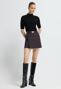 Kvinde i sort kortærmet turtleneck, mørk omslagsnederdel med bælte og knæhøje sorte teksturerede støvler, som står foran en ensfarvet hvid baggrund.