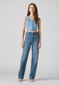 Gilet en denim bleu clair, court, avec des boutons sur le devant et deux poches poitrine, associé à un jean bleu à taille haute et jambes larges. Texture lisse.