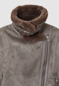 Giacca in suede grigia con colletto in pelliccia marrone e cintura in pelle, caratterizzata da una zip argentata asimmetrica e tasche con zip.