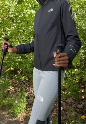 Person iført en sort North Face-jakke og lysegrå North Face-leggings holder sorte vandrestave, mens de vandrer i et grønt, løvfuldt skovområde.