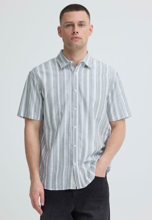 Homme debout devant un fond uni, portant une chemise à manches courtes à rayures verticales gris clair et blanc, boutonnée, et un pantalon noir.