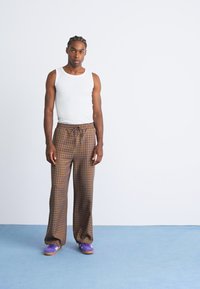 Junger Mann, der auf einem blauen Boden steht, trägt ein weißes ärmelloses Shirt, braune karierte Hosen und lila Sneakers vor einer schlichten weißen Wand.