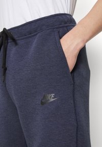 Tumši zilo sporta bikses ar mīkstu tekstūru, elastīgu jostu ar auklu un melnu Nike logo kreisajā augšstilbā.