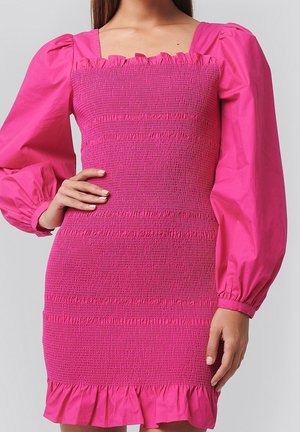 Mujer con vestido fruncido rosa brillante con mangas largas abullonadas y bajo con volantes, posando con una mano en la cadera.