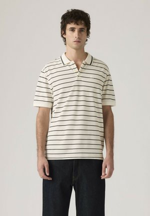 Nuori mies, jolla on valkoinen lyhythihainen poolopaita musta-horisontaalisilla raidoilla ja tummat farkut, seisoo yksinkertaista vaaleaa taustaa vasten.