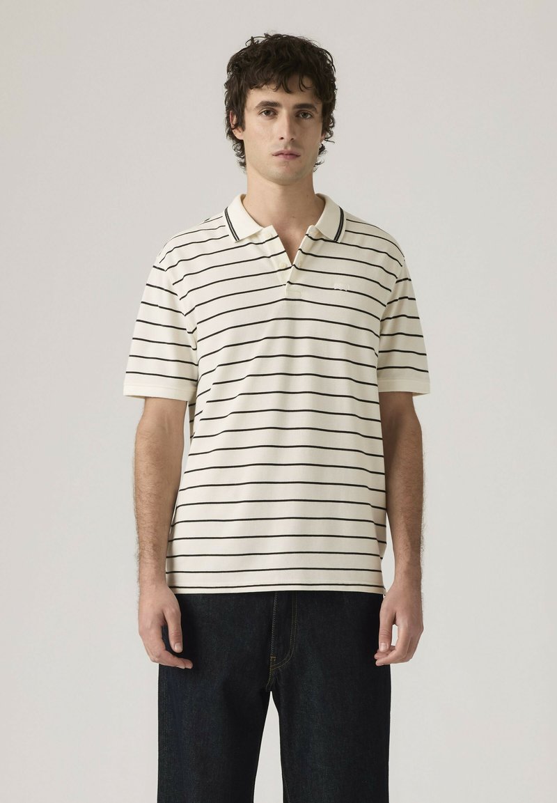 Joven con una camisa tipo polo blanca de manga corta con rayas horizontales negras y jeans oscuros, de pie frente a un fondo claro y liso.